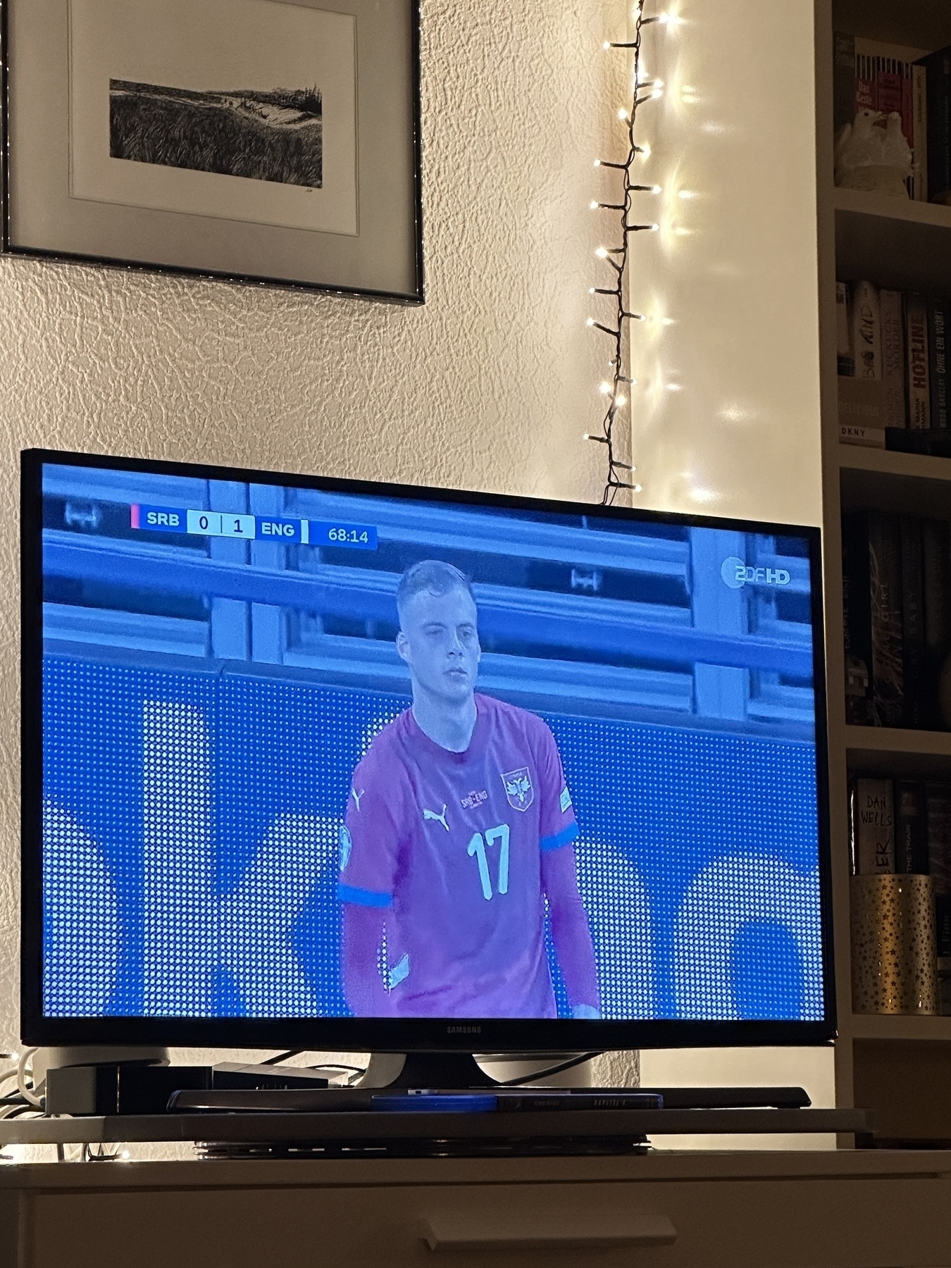 A television screen displays a football match between Serbia and England, with a player in a red jersey and the score at 0-1, surrounded by a cozy living room setting.