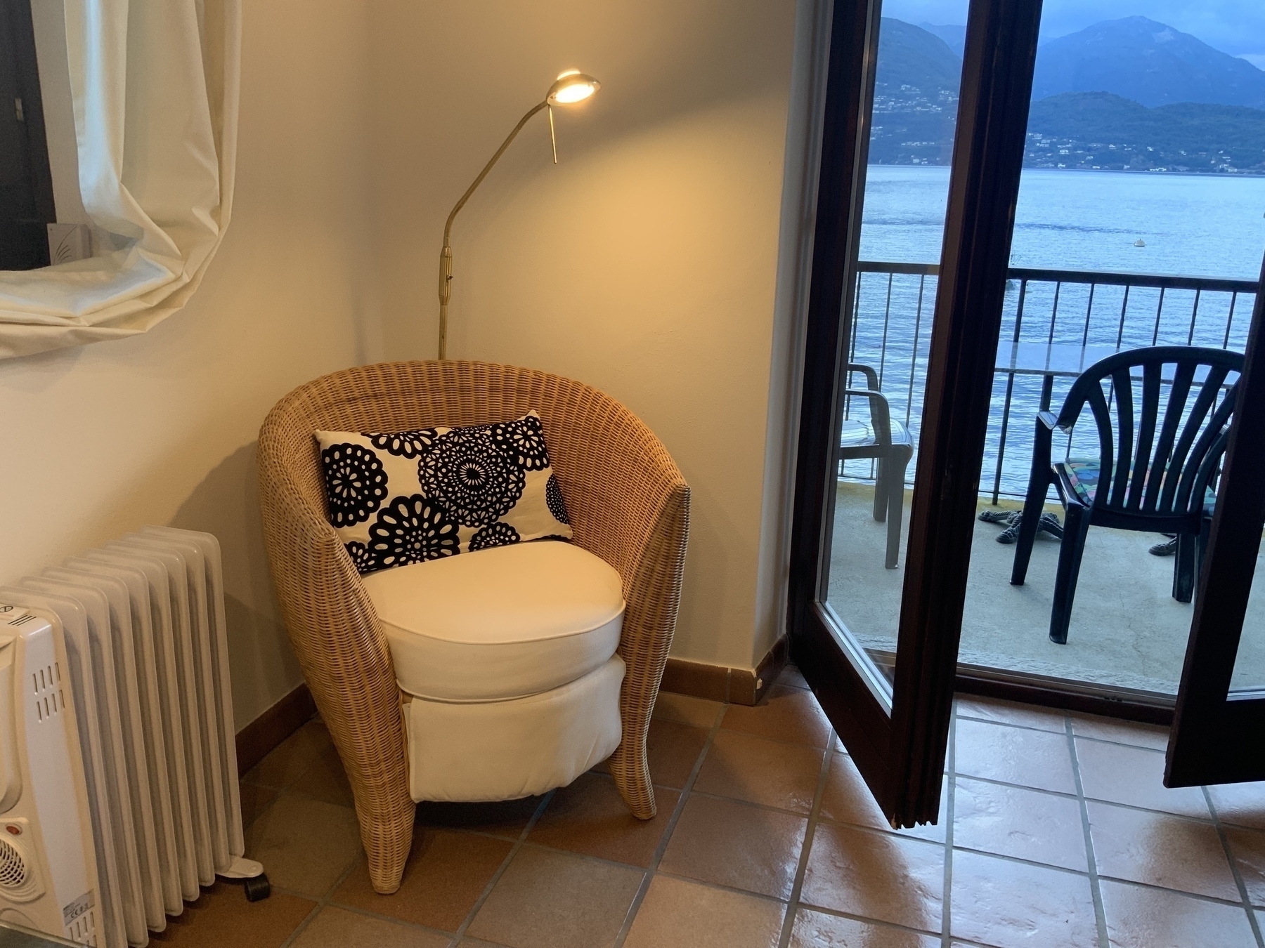 A cozy wicker chair with a black-and-white floral cushion, situated next to a heater and a floor lamp, facing an open door that leads to a balcony with lake and mountain views.